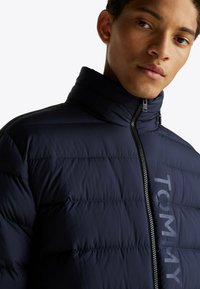 Marine pufferjack met een hoge kraag, voorzien van een horizontale gestikte textuur, ritssluiting en een prominent logo op de voorkant.