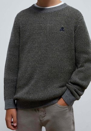 Personne portant un pull gris tricoté texturé avec des poignets et un ourlet côtelés, main partiellement dans la poche d'un pantalon gris clair.