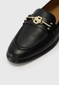 Black leather loafer with a smooth texture, rounded toe, stitched detailing, and a gold-tone logo accent on the front strap.