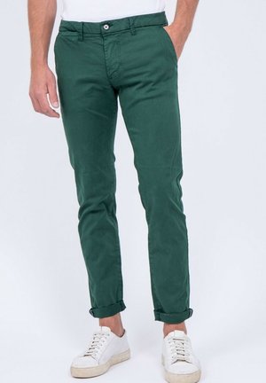 Homme portant un pantalon slim vert foncé retroussé et des baskets blanches, se tenant debout avec une main dans la poche contre un fond uni.