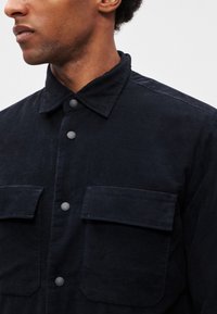 Tamnoplava majica od mekanog tkanja; ima šiljasti ovratnik, sive gumbe i dva džepa na prsima s poklopcima. Teksturirani, ležerni dizajn.