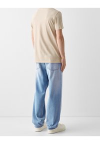Beige cotton t-shirt with short sleeves paired with light blue denim jeans, featuring a relaxed fit and subtle fade at the hips, worn with white shoes.