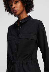 Giubbotto tuta nero con cintura, dotato di colletto, due tasche sul petto, chiusura con bottoni e vita arricciata per una vestibilità regolabile. Tessuto denim liscio.