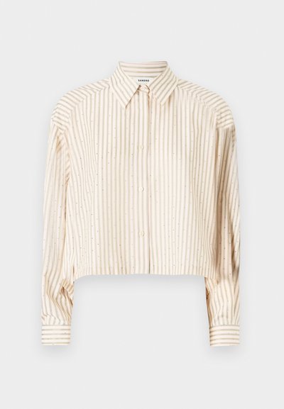 Cremefarvet cropped skjorte med vertikale striber, knaplukning og metalliske prikker på stoffet. Lange ærmer med stribede manchetter.