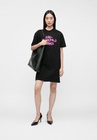 Czarna bawełniana sukienka t-shirtowa z krótkimi rękawami, z różowym logo. Modelka trzyma czarną skórzaną torbę i ma na sobie czarne szpilki.