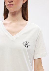 Biele bavlnené tričko s dizajnom V-krku. Na ľavej strane hrudníka má čierne logo Calvin Klein. Hladká textúra a krátke rukávy.
