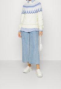 Person wearing a white and blue patterned sweater over a light blue shirt, wide-leg cropped jeans, white platform shoes, and holding a white chain handbag.