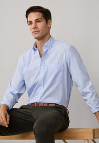 Camicia azzurra chiara con righe bianche verticali, dotata di tasca e dettaglio del logo. Indossata con pantaloni scuri e una cintura marrone.