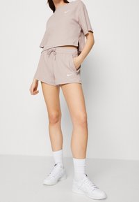 Kvinna i ribbad beige topp med kort ärm och matchande shorts med sidfickor, vita strumpor och vita Nike sneakers, som står mot en vit vägg.