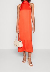 Frau in einem leuchtend orangefarbenen, ärmellosen Halskleid, die eine cremefarbene, gesteppte Handtasche hält und weiße hohe Sandalen trägt.