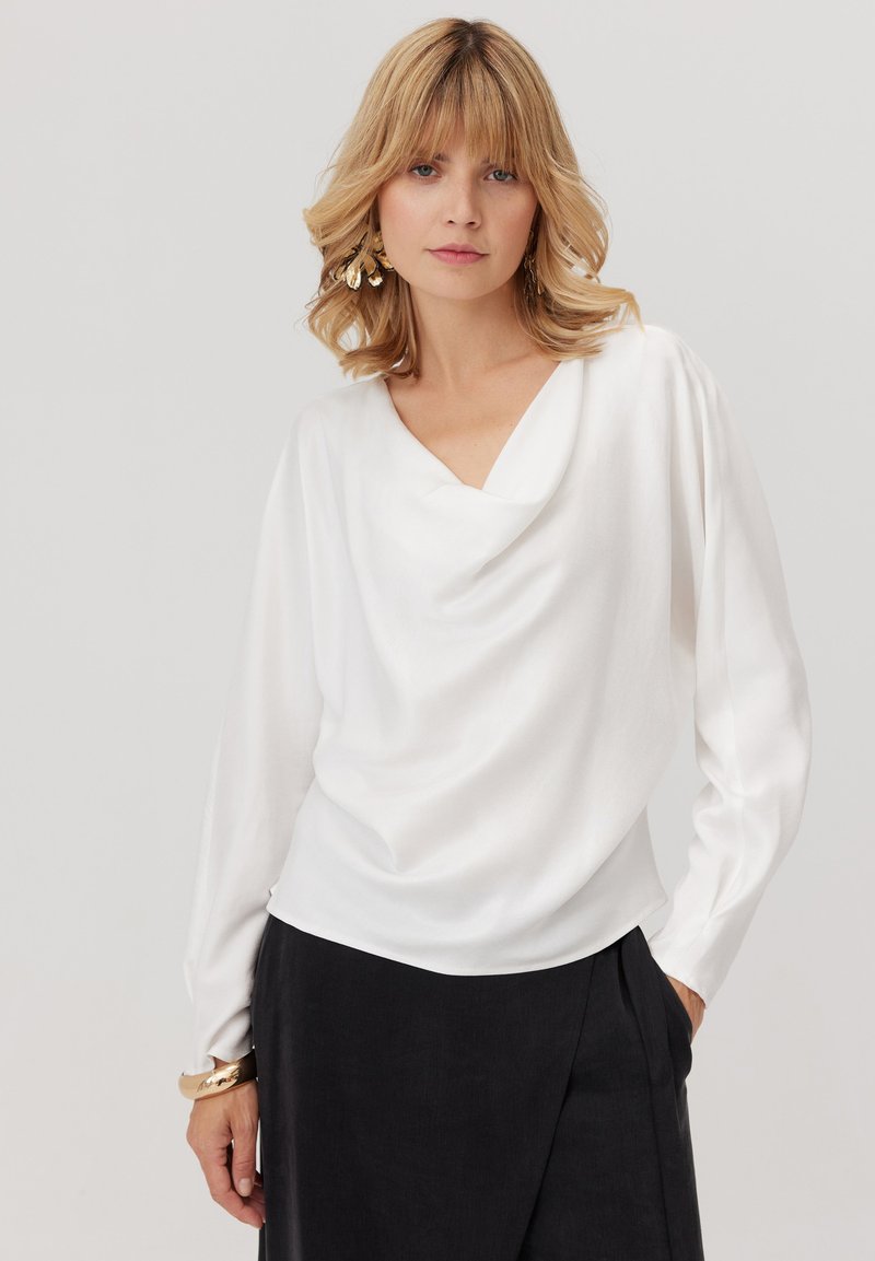 Biała, luźna bluzka z długim rękawem i dekoltem w kształcie litery V, wykonana z gładkiego materiału. Zestawiona z ciemnymi, plisowanymi spodniami. Modelka ma na sobie złote kolczyki w kształcie kół.