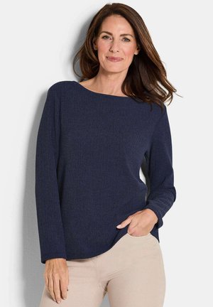 Frau mit langen braunen Haaren, die eine navyblaue Bluse mit langen Ärmeln und beigefarbene Hose trägt, steht mit einer Hand in der Tasche vor einem weißen Hintergrund.