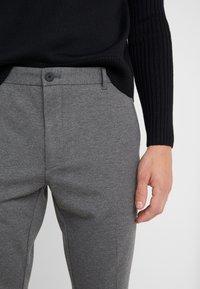 Man in een grijze tailorpantalon en een zwarte ribbeltrui, met de hand rustend naast de dij tegen een effen achtergrond.