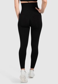 Leggings neri a vita alta realizzati in tessuto elasticizzato, con una texture liscia e un design aderente. Indossati con scarpe da ginnastica bianche.