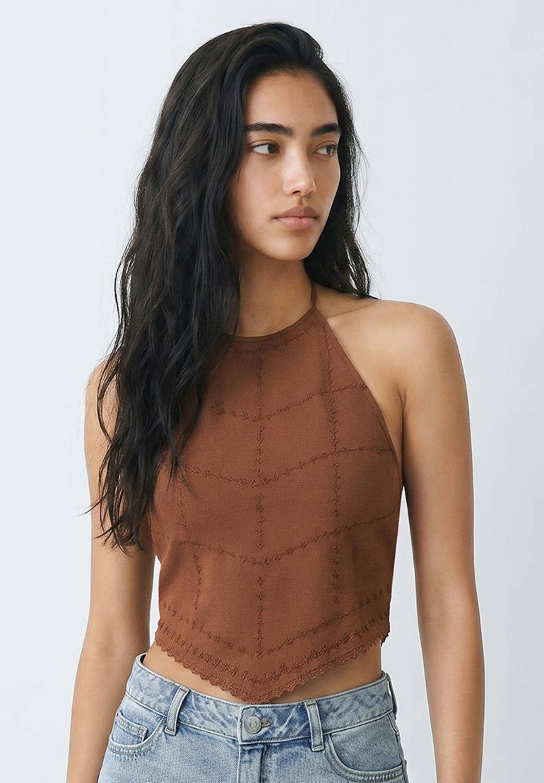 Žena s dlouhými tmavými vlasy, která nosí hnědý bezrukávový halter top a světle modré džíny, se dívá do strany na jednoduchém pozadí.