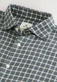 La chemise à carreaux présente un mélange de couleurs bleu, marron et crème, avec un col boutonné et des boutons blancs, fabriquée en tissu doux.