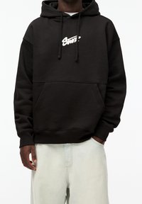 Černá mikina s klokánkovou kapsou, kapucí na stažení a bílým logem na hrudi. Vyrobena z měkkého materiálu s žebrovanými manžetami a lemem.