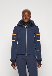 Navy blue puffer jacket with zip closure, hood, and orange, white, and black striped accents on sleeves. Features zippered pockets and logo.