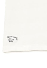 Weißer Stoffecken mit einem kleinen genähten Etikett, das ein Krokodil-Logo und den Text "Authentic Lacoste Est. 1933." zeigt.