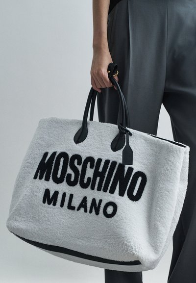 Χέρι που κρατάει μια μεγάλη μαλακή λευκή τσάντα tote με μαύρο κείμενο "MOSCHINO MILANO" και μαύρες λαβές, φορεμένη με σκούρα παντελόνια.