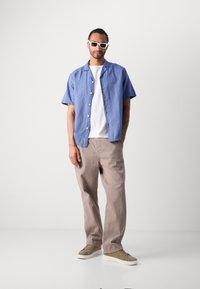 Camicia leggera azzurra a maniche corte con bottoni sopra una maglietta bianca, abbinata a pantaloni larghi beige e sneakers beige. Completato con occhiali da sole.
