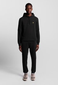 Felpa nera con tasca frontale, cappuccio con cordino e un piccolo logo giallo. Abbinata a pantaloni sportivi neri e scarpe da ginnastica beige.