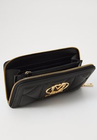 Carteira de couro preto acolchoado com ferragens em tom dourado, apresentando um logo proeminente e vários compartimentos internos, fechada por um zíper.
