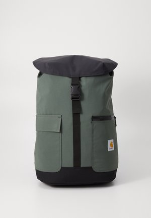 Sac à dos vert et noir avec rabat supérieur, sangle centrale à boucle, deux poches avant et un petit patch logo Carhartt sur la poche inférieure droite.