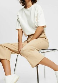 Personne assise sur une chaise transparente, portant un t-shirt blanc ample, un pantalon beige coupé et des baskets blanches sur un fond uni.