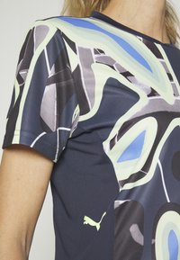 Close-up van een persoon die een sportshirt met korte mouwen draagt met abstracte blauwe, grijze en witte patronen en een klein Puma-logo vlak bij de taille.