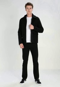 Jachetă neagră cu fermoar și guler, purtată peste un tricou alb. Pantaloni negri și adidași negri cu accente albe completează ținuta.