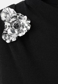 Broche floral plateada sobre un suéter negro texturizado. La broche cuenta con pétalos superpuestos y un detalle de diseño central, realzando la apariencia de la prenda.