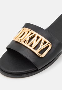 Sandale à glissière noire avec une large bride ornée d'une plaque logo DKNY en métal doré sur le dessus.