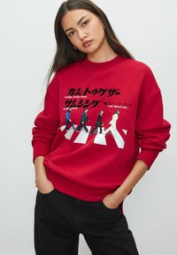Punane sweatshirt, millel on graafiline trükk, mis on inspireeritud The Beatles'ist, valge teksti ja tänava ületamise disainiga. Kootud mansetid ja äär.