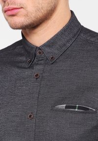 Mørkegrå skjorte med knapper og tekstureret stof, der har en lille lomme med mønstret foring og brune knapper ved kraven.