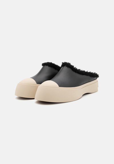Chaussures noires à enfiler avec une bordure en fausse fourrure et des orteils ronds en caoutchouc blanc, dotées d'une semelle plateforme chunky beige. Fini mat lisse.