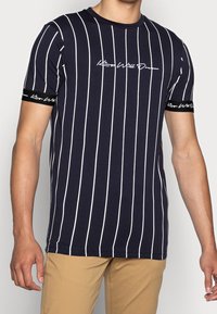 Marinblå t-shirt med kort ärm och vita vertikala ränder, med en skriptlogotyp och kontrasterande svarta band på ärmarna. Tillverkad av bomull.