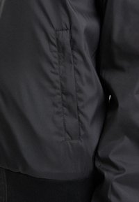 Veste en tissu noir avec une poche fente verticale et une ceinture côtelée, montrant une texture légèrement froissée et des détails de couture.