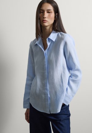 Junge Frau trägt ein hellblaues, langärmliges Hemd mit Knöpfen und dunkle Jeans, sie steht mit einer Hand in der Tasche vor einem schlichten Hintergrund.