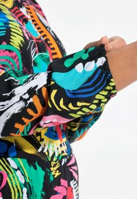 Bunt gemustertes Stoffdesign mit schwarzem Grund, das lebhafte Wirbel und Formen in Pink, Grün, Orange und Blau zeigt. Elastische Bündchen sind sichtbar.