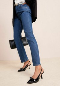 Niebieskie dżinsy z wysokim stanem w połączeniu z czarnymi lakierowanymi czółenkami na obcasie z paskiem z tyłu i kokardą, uzupełnione czarną torebką.