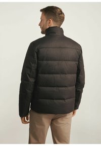 Czarna kurtka puchowa z wysokim kołnierzem, przeszywanymi panelami i matową fakturą, w połączeniu z jasnobeżowymi spodniami. Brak widocznych znaków tożsamości czy akcentów.