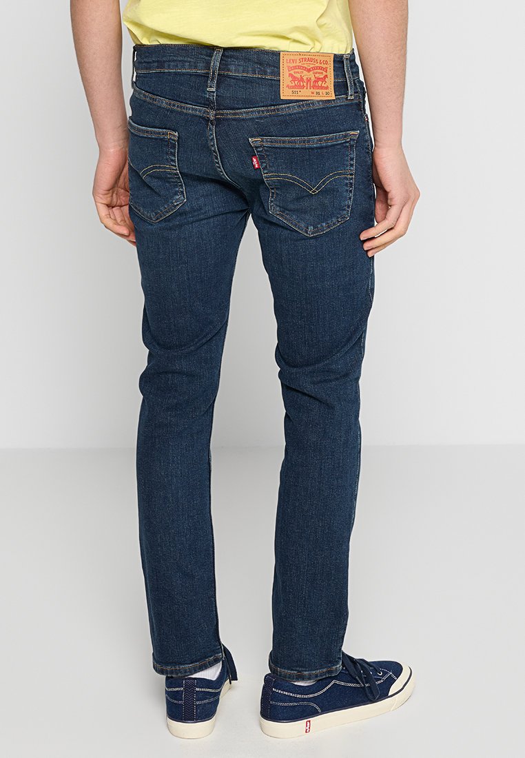 Dunkelblaue Jeans mit schmaler Passform, mit einem Etikett auf der Rückseite und zwei Taschen. Der Stoff erscheint glatt mit einem leichten Glanz.
