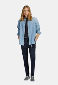 Camicia di jeans azzurro chiaro sopra una maglietta nera, abbinata a pantaloni blu scuro e sneakers grigie. Fit dritto, con maniche arrotolate e dettagli con bottoni.