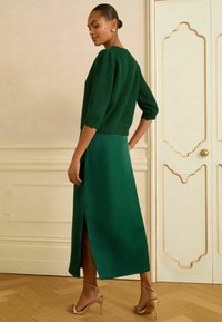 Pulover verde tricotat cu mâneci trei sferturi, asortat cu o fustă lungă, netedă, din satin verde, având o fanta laterală. Sandalele aurii cu bretele completează ținuta.