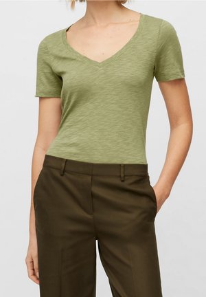 Frau trägt ein hellgrünes T-Shirt mit V-Ausschnitt und kurzen Ärmeln, das in dunkelolivfarbene, maßgeschneiderte Hosen gesteckt ist, mit den Händen in den Taschen.