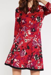Robe fleurie rouge à manches longues ; présente une jupe plissée et une bordure noire. Le tissu a une texture lisse avec un motif vibrant de roses.