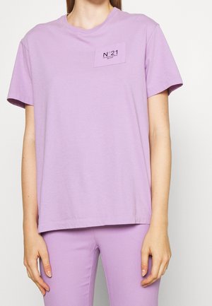 Osoba ubrana w lawendową koszulkę z krótkim rękawem i pasujące spodnie, z małą prostokątną etykietą na piersi z napisem "N°21 VIA SANTO SPIRITO MILANO".