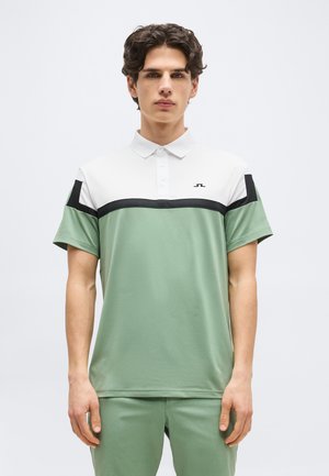 Νεαρός άντρας που φοράει πόλο μπλουζάκι με κοντά μανίκια, σε λευκά, μαύρα και απαλό πράσινο χρωματικά μπλοκ, στέκεται μπροστά σε ένα απλό φόντο.