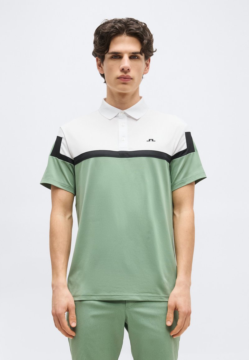 Νεαρός άντρας που φοράει πόλο μπλουζάκι με κοντά μανίκια, σε λευκά, μαύρα και απαλό πράσινο χρωματικά μπλοκ, στέκεται μπροστά σε ένα απλό φόντο.